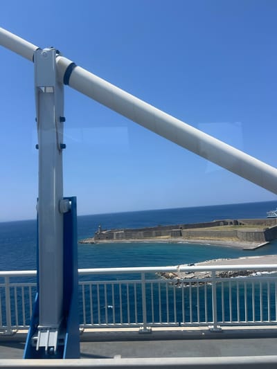 The image captures a view from what appears to be a bridge or ferry, looking out over the sea towards a fortified coastline. In the foreground, large white and blue metal structural elements dominate the left side of the frame, suggesting the vantage point is on a man-made structure. A white railing with vertical bars runs horizontally across the bottom of the image, separating the viewer from the sea.
The sea itself is a deep blue, with visible ripples and a lighter turquoise hue near the shore. On the coastline, a substantial stone fortress with a lighthouse stands prominently. The fortress appears to be old, with a weathered stone construction and arched openings visible. A small beach area is situated in front of the fortress, with some rocky outcrops and what might be a small boat or debris.
Further in the distance, a modern white boat with a blue stripe is visible in the water, suggesting ongoing activity in the area. The sky is a clear, bright blue. The overall scene suggests a sunny day in a coastal location. This view is consistent with the location in the Municipal Unit of Antirrio, Greece, which is known for its bridge and surrounding historical fortifications.