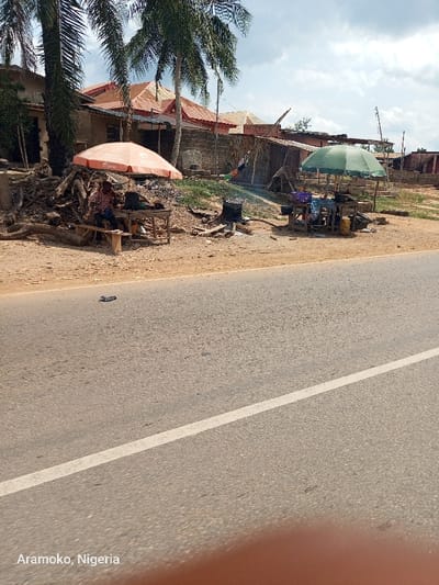 The image shows a paved road with a white stripe running down the center, located in Okinni, Nigeria (latitude: 7.832, longitude: 4.513). There are two open-air stalls on the right side of the road, both with umbrellas. One stall has an orange umbrella and is attended by a person sitting on a stool, and the other stall has a green umbrella and is unattended. The stalls are located on a dirt shoulder along the side of the paved road. Behind the stalls are a few buildings with tin roofs. The image is taken from the perspective of a vehicle driving on the road. The image is blurry and the quality is poor.