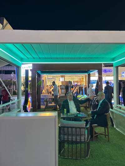 The image depicts an outdoor seating area with artificial turf flooring, illuminated by a vibrant green light emanating from the ceiling structure. Two men in suits are seated at a table, engaged in conversation, with one gesturing. The background reveals a commercial setting with storefronts and people moving about. One storefront displays a sign with "o George" in blue lettering. The overall scene suggests a public gathering or event in an urban environment. The location is identified as Al-Ghizlaniya, Syria.