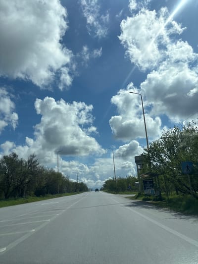 The image depicts an open, two-lane asphalt road under a bright blue sky filled with large, scattered cumulus clouds, with a visible sun flare in the upper right quadrant. This scene is located in the city of Drama, Greece.
The road extends straight into the distance, with a slight uphill grade. White solid lines mark the road edges, and faded white dashed lines are visible in the center, indicating two-way traffic. Faint, worn white markings on the left lane are present. Along both sides of the road, green grass and rows of deciduous trees with nascent spring foliage are visible.
On the left side of the road, multiple tall, slender streetlights with dark fixtures and reddish-brown poles are spaced out. On the right side, closer to the foreground, a similar streetlight stands near a bus stop shelter with a light-colored canopy and a visible advertisement poster featuring the word "PATERIS" and "BAKERY" in Greek. Behind the bus stop, a tall, grey, concrete water tower structure is present. A blue rectangular sign with a white pedestrian symbol is mounted near the bus stop. Utility poles with overhead wires are also visible on the right.
In the far distance, a single dark-colored passenger vehicle is observable on the road, traveling away from the camera. There are no other visible people or active interactions in the scene. The perspective suggests the image was captured from inside a vehicle, looking forward.