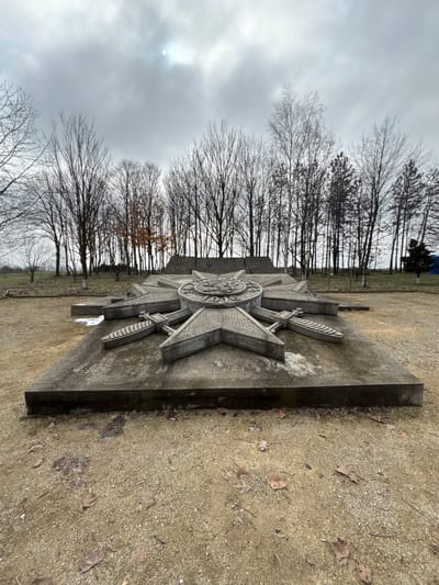 A large concrete monument, identified as being in Shumentsi, Bulgaria, is centered in the foreground of an outdoor scene under an overcast sky. The monument consists of a prominent eight-pointed star design, approximately 1.5 to 2 meters in diameter, elevated on a rectangular concrete base. The central medallion of the star features an embossed emblem with the year "1916" clearly visible, flanked by what appear to be depictions of stylized swords or daggers between some of the star's points. The concrete exhibits weathering and minor discoloration. Behind the primary star structure, additional lower-profile concrete elements form part of the monument. The ground surrounding the monument is a light brown, sandy or gravelly surface, sparsely covered with dry, fallen leaves. In the mid-ground and background, a dense line of tall, bare deciduous trees, indicative of winter, forms a natural barrier. A few evergreen trees are discernible on the right side of the background. The sky is uniformly gray and heavily clouded, with the sun's diffuse light faintly visible through the cloud cover at the top center. No individuals or active subjects are present in the image.