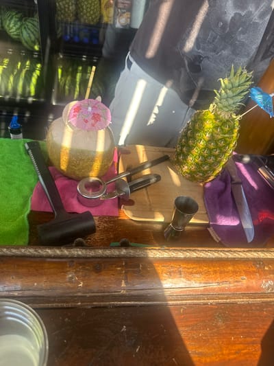 In Protaras, Cyprus, on a sunlit day, a scene unfolds at what appears to be an outdoor bar or fruit stand. A whole coconut, filled with a light-colored liquid and adorned with a pink floral umbrella, sits next to a whole pineapple, also decorated with a blue umbrella. Both are positioned on a wooden counter.
To the left, a vibrant green towel is spread out, partially covered by a black mallet. A pink towel lies beneath the coconut and a chrome-plated tool, possibly an ice scoop. A wooden cutting board is visible behind these items, featuring a black knife and another tool with a black handle.
To the right of the pineapple, a purple towel is draped, with a silver knife resting on it. A small silver jigger, used for measuring liquids, is positioned in front of the cutting board. In the background, a display refrigerator is filled with watermelons and pineapples, suggesting a focus on fresh fruit. The overall atmosphere is one of tropical refreshment, enhanced by the bright sunlight casting shadows across the wooden counter.