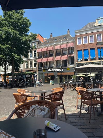 The image captures a bustling outdoor seating area of cafes and restaurants on a sunny day in the city of Zwolle, Netherlands. In the background, a McDonald's restaurant is prominently visible with its iconic golden arches. Adjacent to it are other establishments with outdoor seating, featuring umbrellas and tables. Several people are seen dining and socializing at these tables. Two bicycles are parked near the McDonald's. The ground is paved with cobblestones, and a large green tree provides shade in the left portion of the frame. The architecture of the buildings suggests a historic European city setting, with multi-story buildings featuring traditional window designs and awnings. The scene conveys a lively atmosphere typical of a public square or marketplace during pleasant weather.