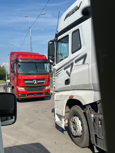 The image shows two semi-trailer trucks on a paved surface, likely a road or parking lot, under a clear blue sky with some clouds. The truck on the left is red and bears a Mercedes-Benz emblem on its grille. The truck on the right is white and has "FAW" written on its cab. Both trucks appear to be parked or stopped. The environment suggests a sunny day, possibly in a rural or semi-urban area, given the presence of trees in the background and a lamppost. There are no visible people in the immediate vicinity of the trucks, though a reflection in the side mirror of a vehicle in the foreground suggests a driver may be present in that vehicle. The image is framed from a low angle, looking up at the trucks. There is no specific indication of the city of Verkh-Sosnova, Russia, within the image itself.