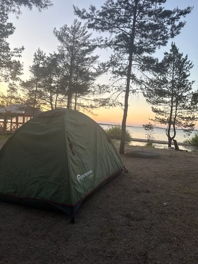 A serene outdoor scene captures a camping setup by a tranquil body of water, likely a lake, during either sunrise or sunset. The image, set in Zerendi, Kazakhstan, is dominated by a dark green camping tent positioned in the foreground and mid-ground on the right side. The tent, branded "OUTVENTURE COVE 2", faces towards the water.
In the mid-ground and background, tall pine trees with dark, silhouetted branches frame the view, their needles visible against the sky. Between the trees and beyond the tent, a calm body of water stretches across the horizon, reflecting the warm hues of the sky. The sky transitions from a soft orange and yellow glow near the horizon, typical of dawn or dusk, to a pale blue overhead.
The ground in the foreground is a mix of dark earth, gravel, and sparse vegetation, consistent with a natural campsite. A small, dark rock is visible near the water's edge on the right. Further into the background, partially obscured by the tent and trees, a wooden structure or pier can be faintly discerned along the shoreline. The overall atmosphere is one of peaceful wilderness, suggesting a quiet moment in nature.