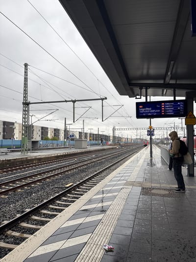 The image depicts a train station platform in Bad Vilbel, Germany, under an overcast sky. The foreground shows a wet platform paved with dark grey square tiles and interspersed white and dark grey striped tactile paving. Near the platform edge, a crumpled red and white cigarette packet lies on the ground. A circular metal manhole cover is visible further back on the platform.
On the right, beneath a dark grey overhead canopy structure, a person wearing a tan/khaki hooded jacket, dark pants, and dark shoes stands facing away from the camera, looking towards the tracks. They appear to be holding a small electronic device. A light-colored bag is slung over their left shoulder. Partially visible behind them are the lower legs and feet of another individual. Above them, a digital display screen hangs from the canopy, showing train schedules for the S6 line, with destinations including Langen (Hess) and Friedberg (Hess), and corresponding wait times. Warning signs, including a yellow triangle with an exclamation mark, are also present.
To the left of the platform, multiple parallel train tracks stretch into the distance. The tracks consist of metal rails set on concrete sleepers embedded in dark grey ballast. Overhead catenary wires and their elaborate support structures, including tall metal lattice poles, span across the tracks. In the far distance, a red train or locomotive is visible on the tracks.
Across the tracks, a second platform is visible, featuring concrete shelters and benches. A blue sound barrier or fence runs along the far side of this platform, behind which multi-story residential or commercial buildings with facades of varying dark and light shades are present. Small "Bad Vilbel" station signs are visible on both platforms.