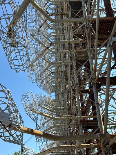 This image depicts the abandoned Duga radar complex, located near Chernobyl, Ukraine. The photograph, taken from a low angle looking upwards, showcases the immense scale of the Soviet-era radar system against a clear blue sky. The structure is a massive array of metal beams, pipes, and wire mesh, painted a faded off-white and showing signs of rust and wear. Various sections of the radar, including what appear to be antenna elements, are visible, stretching into the distance. The intricate network of supports and interconnected components suggests a complex technological installation. The scene is devoid of human presence and any signs of recent activity, emphasizing the derelict and abandoned nature of the site. In the background, glimpses of green foliage can be seen, indicating the surrounding natural environment.