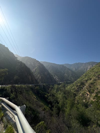 This photograph captures a wide, expansive view of a mountainous canyon landscape within the Unincorporated Santa Monica Mountains, United States of America, under a clear, bright blue sky.
The scene is dominated by a deep valley carved between steep, heavily vegetated mountainsides. The slopes are covered in a mix of dense green foliage, likely trees and shrubs, with visible rocky outcrops and some areas of drier, darker, possibly burnt vegetation, particularly in the foreground and lower left. Atmospheric haze is present between the distant mountain peaks.
A two-lane paved road or highway snakes along the contours of the left mountainside, approximately midway up its slope. Several small vehicles, including at least one white and one dark-colored car, are visible traveling on this road. A bridge structure supports a segment of the road in the mid-ground. Overhead utility power lines extend diagonally across the upper left quadrant of the image, running parallel to the left mountainside.
In the immediate foreground, a gray metal guardrail, constructed from two parallel corrugated beams supported by vertical posts, extends from the bottom left corner towards the center. Below the guardrail, the terrain drops steeply into the canyon, covered in dry grasses and dark, sparse brush.
The sky occupies the upper two-thirds of the image, a vivid blue. A bright sun flare emanates from the upper left, indicating direct sunlight. A faint, white contrail from an aircraft is visible as a thin horizontal line in the sky above the left mountain ridge. No individuals are discernible within the frame.