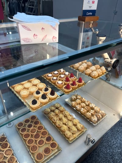 The image shows a display case in a bakery or pastry shop in Boca del Río, Mexico. Inside the glass case are trays of various small tarts. One tray features tarts with a meringue swirl. Another has tarts filled with chocolate and topped with fresh raspberries. A third contains tarts with white cream and blackberries. There is also a tray of pecan tarts and another with chocolate tarts. Additionally, there is an iZettle card reader attached to the glass. To the left of the pastry display case is a tissue dispenser. The background suggests an urban environment with reflections of buildings visible on the glass.
