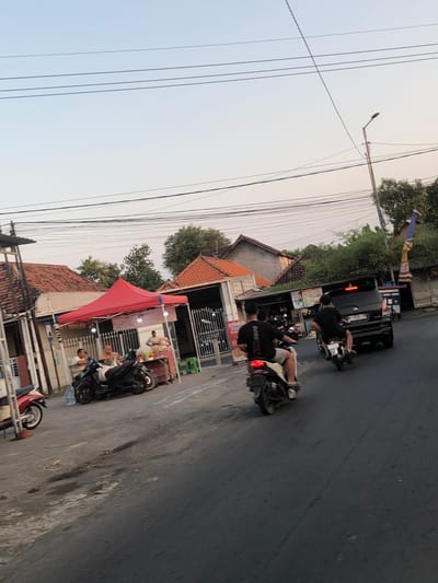 This is a full, eye-level, outdoor shot captured in Glagah Arum, Indonesia, at dusk. The scene depicts a street with several buildings lining one side, featuring traditional red-tiled roofs. In the foreground, the asphalt road is visible, with various motorcycles parked and in motion.
On the left side of the street, a red tent is set up, beneath which several people are seated at a makeshift food stall. A motorcycle is parked in front of the tent. Further down the street, a dark-colored van is visible, and two men on motorcycles are riding past it. One of the motorcycles is in the immediate foreground, traveling away from the viewer.
The environment appears to be a local commercial area or a roadside market. Overhead, a network of power lines crisscrosses the sky, with a street lamp visible on the right. The sky is clear and exhibits the soft hues of twilight. The overall impression is that of a casual, active street scene in a residential or commercial district.
