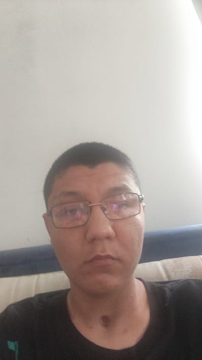 A close-up, low-angle selfie of a person wearing glasses and a black t-shirt. The person has short, dark hair and a light complexion. There is a dark mark on their neck, possibly a birthmark or injury. The background appears to be a room with a light-colored wall and a dark blue couch. There are no discernible landmarks or contextual clues to indicate the location as Становая, Russia.