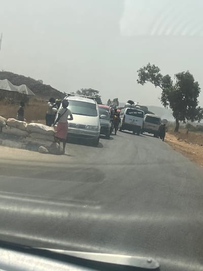 This image depicts a roadside scene in Assob Hausa, Nigeria, viewed from within a vehicle. The foreground shows a paved road, where a line of vehicles appears to be stopped or moving slowly.
On the left side of the road, a stack of approximately six large, light-colored sacks, appearing to contain granular material, is arranged. Several individuals are present in this area. One woman, wearing a red skirt and a white top with a dark shoulder bag, stands with her back to the camera, positioned between the sacks and a silver Volkswagen minivan. Another individual with a yellow head covering is partially visible further left, seemingly interacting with another person.
The silver Volkswagen minivan, equipped with a roof rack carrying various items, is stationary on the road. Directly behind it is a dark-colored sedan. Further down the road, a white minivan with a loaded roof rack is visible, displaying "LET ME TRY" in black text on its rear window. Another light-colored van is positioned behind it. Multiple individuals are observed walking between and alongside these vehicles on the road.
The right side of the road features a dry, unpaved shoulder of brown earth. A single tree with sparse green foliage stands on this shoulder. The background on the left reveals a dry, hilly landscape with minimal vegetation and several white, tent-like structures. The sky is uniformly hazy and light grey-blue.