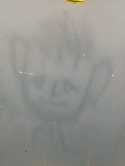 The image shows a rough, gray surface, possibly a wall or a cabinet. There is a faint, smudged image of a face or figure visible on the surface, appearing to be a shadow or a stain. The texture of the surface is granular. At the top of the image, a yellow and black label with some text is partially visible. The image appears to be an interior shot, given the lighting and lack of outdoor elements. There are no people or animals present in the frame. The exact location within Bremerhaven, Germany cannot be determined from the image.
