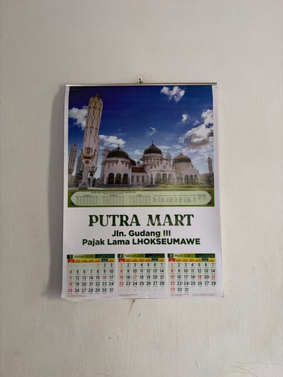 The scene depicts a commercial wall calendar for the year 2026, affixed to a plain, off-white wall in the city of Uleemeuria, Indonesia. The calendar is secured near its top edge by a single, dark-colored tack or small nail.
The top portion of the calendar features a wide-angle color photograph of a large, white mosque with multiple dark domes and several distinctive, tall, white, umbrella-like structures. The mosque is set against a bright blue sky with scattered white clouds. Below the image, prominent green text identifies "PUTRA MART" with the address "Jln. Gudang III" and "Pajak Lama LHOKSEUMAWE". A narrower green strip beneath this text contains smaller white text indicating prayer schedules: "JADWAL WAKTU SHOLAT UNTUK KOTA JAKARTA MENURUT KEMENAG RI" and "JADWAL WAKTU SHOLAT DARUSSALAM." The bottom section of the calendar displays three monthly grids: "1 Januari 2026," "2 Februari 2026," and "3 Maret 2026." Sundays are highlighted in red within these grids. The surrounding wall exhibits minor textural variations and faint vertical markings below the calendar. No individuals or other objects are present in the visible frame.