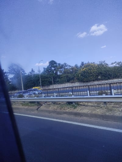 A view from a moving vehicle shows a multi-lane highway separated by a metal barrier. Beyond the barrier, a higher road is visible with cars and possibly a yellow bus. Trees line the horizon with a blue sky overhead. A lone cloud floats in the distance. The road is dark, and the sky is bright. The perspective is low, capturing the road ahead and the scenery passing by.