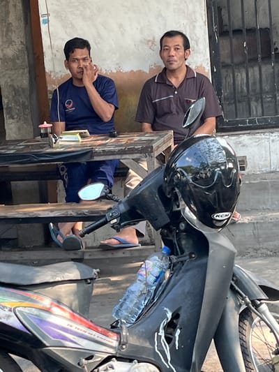 In Kludan, Indonesia, two men are seated at a wooden table outdoors, with a motorcycle in the foreground. The man on the left, wearing a navy blue t-shirt with a logo, is touching his nose with his right hand. The man on the right, in a brown polo shirt, looks towards the viewer. A black motorcycle helmet rests on the motorcycle's front. The environment appears to be a street or an open-air seating area, with a weathered wall and barred windows visible in the background.