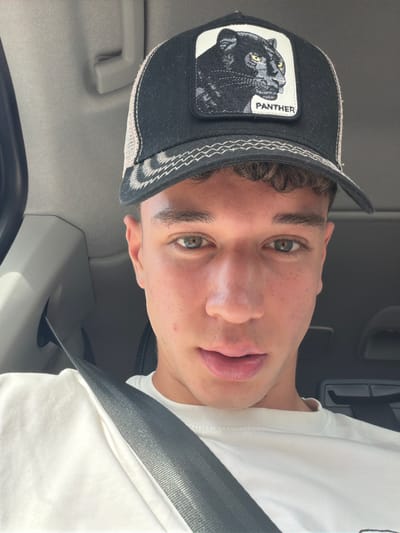 A young man is shown in a close-up selfie, likely taken inside a vehicle due to the presence of a seatbelt and interior car elements. He is wearing a white shirt and a black baseball cap with a mesh back. The cap features a patch depicting a black panther with the word "PANTHER" embroidered next to it. The man has short brown hair, fair skin, and light-colored eyes. He is looking directly at the camera with a neutral expression. The car's interior is light gray, and a portion of the steering wheel is visible. The location in the city of Rivoli, Italy is not discernible from the image provided.