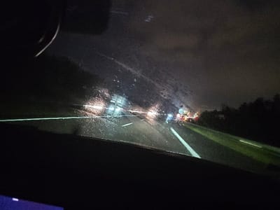 The image depicts a nighttime scene from inside a car driving on a highway. Rain streaks the windshield, blurring the lights of other vehicles ahead. The road is visible, with white lane markings. Trees line the side of the highway. The dashboard is dark. The sky is overcast. The visibility is poor.