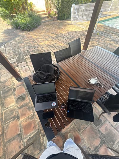 This is an outdoor scene likely in a residential area of Urbanització La Canyada, Spain, evidenced by the Spanish-style tiled patio. The shot is taken from a high angle, looking down at a wooden outdoor table set on the patio. Two laptops are on the table, one black and one silver, along with a black backpack, a smartphone, a glass, and a white charger with a cable. A person is partially visible, wearing a light blue and white striped shirt and tan shoes. The table is surrounded by black wicker chairs. In the background, there's a glimpse of greenery and a white metal structure. The lighting suggests it is daytime.