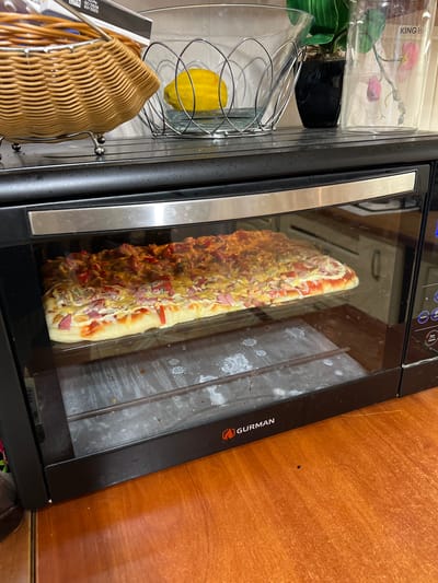 The image shows a long, narrow pizza with various toppings, including cheese, tomatoes, and what appears to be ham, being cooked inside a black oven with a glass door. The pizza is positioned lengthwise inside the oven.
To the left of the oven, there are several kitchen items visible. These include a clear plastic container with what looks like artificial flowers and a black pot, and a silver wire rack holding a yellow lemon. Below that, a woven basket is partially visible, and a box with a barcode and text is also present.
The oven has a sleek design with a stainless steel handle on the left. The control panel of the oven is visible at the top, displaying a digital interface with buttons. The brand "GURMAN" is visible in orange lettering on the right side of the oven's front panel.
The background to the right of the oven is a wooden surface, likely a kitchen cabinet or countertop. The lighting suggests it is daytime. The overall scene is a domestic kitchen setting, and the activity is the preparation or cooking of food. There is some smudging and residue visible on the glass of the oven door, indicating it has been used. The visible text on the box is a barcode and the text "02/2025", likely an expiration date. The text "KING H" is visible on the clear plastic container.