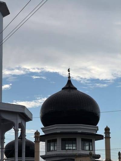 This image depicts a detailed view of a mosque in Ietarek Dua, Indonesia, set against a partly cloudy daytime sky. The central feature is a large, dark, segmented dome, surmounted by a gold-toned finial consisting of a crescent moon and star. This dome rests upon a white, multi-faceted structure that includes rectangular windows. To the immediate left, a portion of another white building, likely an extension of the mosque, is visible with an arched architectural detail and supporting columns. A smaller, dark dome is partially obscured behind this structure. To the right of the prominent dome, a slender, light-colored minaret or decorative spire with an ornate, multi-tiered cap is present. Multiple dark utility lines traverse the upper and middle sections of the frame. No individuals, specific actions, or interactions are visible in the scene. The sky displays a mix of blue patches and white to light grey clouds.