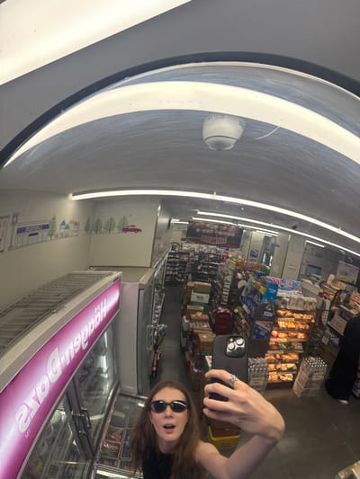 The image depicts a wide-angle selfie taken by a woman reflected in a large, curved mirror within a convenience store. The mirror's curvature causes significant optical distortion, resulting in an arched ceiling and bowed structural elements throughout the scene. The store is located in Changning District, China.
The main subject, a young woman with long, light brown hair, is positioned in the lower right portion of the image. She is wearing dark, oval-shaped sunglasses and a dark top, with her mouth slightly open. She holds a dark-colored smartphone, featuring a prominent triple-lens camera module, in her left hand, using it to capture the image. A silver-colored ring with an intricate, openwork design is visible on her left ring finger.
To the left of the woman, a tall, illuminated Haagen-Dazs freezer unit extends into the background. Its pink display panel shows the brand name "Haagen-Dazs" in mirrored text (appearing as "szaD neggeH") followed by "MT" at the bottom. The freezer shelves hold various ice cream products. Above the freezer, a metallic, gridded surface is visible.
The back wall of the store features a light-colored mural with stylized line drawings of a townscape, including buildings, trees, and a red car. One of the buildings clearly displays the mirrored "NOSWAL" text, indicating a Lawson convenience store. The store's ceiling, distorted into an arch by the mirror, is fitted with multiple linear fluorescent light fixtures. A white, dome-shaped CCTV security camera is centrally mounted on the ceiling.
To the right, aisles of shelving are visible, densely stocked with a variety of convenience store products, including packaged goods, beverages (such as bottled water), and a display of baked goods on tiered racks. In the far-right background, a person wearing dark clothing and white shoes is partially visible, standing near a stack of bottled water cases. The floor is a dark, reflective surface.