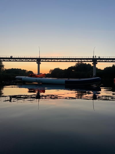 An overhead view captures a serene river scene at dusk in Barysaw, Belarus, with a bridge spanning the water. On the bridge, several figures are silhouetted against the twilight sky. Below, a paddleboard floats on the calm surface of the river, reflecting the bridge and the warm hues of the sunset. Trees line the riverbank, their dark silhouettes contrasting with the vibrant sky. The overall atmosphere is peaceful and contemplative.