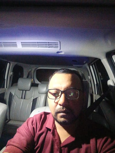 A selfie taken inside a car at night. The subject, a man wearing glasses and a maroon collared shirt, is the primary focus. Behind him, the interior of the car is visible, including gray leather seats and a ceiling air vent illuminated by purple light. The background is dark, suggesting it's nighttime. The man's facial expression is neutral. The image appears to be taken in Awegeutahpaya, Indonesia.