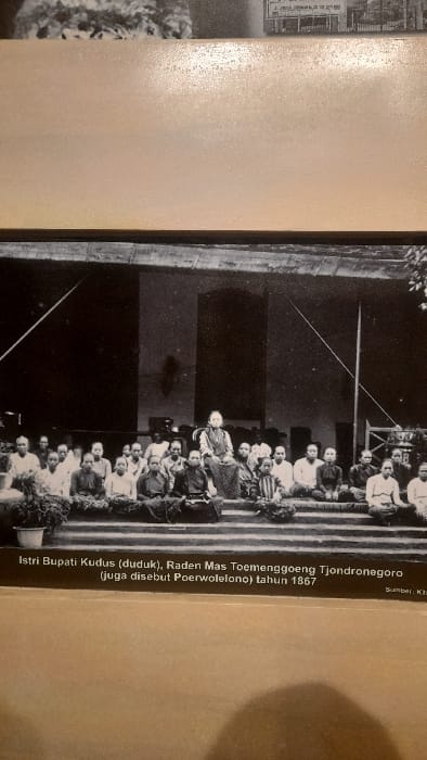 This black and white historical photograph, dated 1867, captures a posed group portrait of approximately 25 to 30 individuals, predominantly women, seated on the steps and ground of a traditional Javanese building. The central figure, identified by the accompanying text as the wife of Raden Mas Toemenggoeng Tjondronegoro (also known as Poerwolelono), the Regent of Kudus, is seated on a distinct chair in the front row, elevated above the others. All individuals are dressed in traditional Indonesian attire, including sarongs and kebayas or similar garments, with their hair typically pulled back.
The scene is set outdoors or within an open-air pavilion (pendopo), with the dark, solid structure of a building forming the background. Visible environmental elements include a potted plant on the left foreground, a tall pole (possibly a flagpole) on the right, and a decorative urn or stand to its right. The image itself appears to be a framed display piece, exhibiting signs of light reflection and texture from a surrounding surface, consistent with being mounted in a museum or exhibition. The depicted event is associated with Kudus, Indonesia, while the physical display of this image is located in the city of Glantengan, Indonesia.