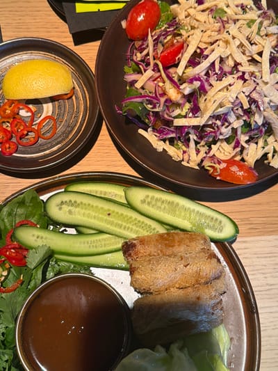 The image displays a meal set on a light-colored tabletop in a restaurant located in Kiryat Ono, Israel. No individuals are visible. The scene features three distinct plates of food. In the upper left, a small, dark ceramic plate holds a yellow lemon half and several thin, circular slices of red chili peppers. To its right, a larger, dark ceramic bowl contains a salad consisting of shredded red cabbage, green leafy vegetables, multiple red cherry tomato halves, black sesame seeds, and crispy yellow tortilla-like strips. In the lower portion of the image, a large, dark ceramic plate with a lighter rim presents a variety of items: several long, sliced green cucumbers, a mound of fresh green herbs (likely cilantro and basil) intermixed with small red chili pieces, a small silver bowl filled with a dark brown dipping sauce, and three golden-brown, cylindrical fried spring rolls or similar items stacked atop light green lettuce leaves. A dark napkin or menu is partially visible in the distant background.