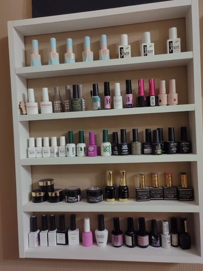 The image shows a white, multi-shelf unit containing an assortment of nail polish bottles and jars. The shelves are filled with rows of bottles of various sizes, shapes, and colors, predominantly displaying shades of pink, white, black, and lighter pastel tones. Some bottles have distinct branding, while others are unbranded or have less legible labels.
The nail polish bottles are arranged neatly in rows on each shelf. The top shelf features bottles with pastel blue and peach colored tops. The second shelf from the top has white bottles with black text and some pink-capped bottles. The third shelf from the top contains a variety of bottles with colored caps and some clear bottles with colored polish. The fourth shelf is populated with mostly black bottles with gold caps, and some jars. The fifth shelf from the top has several black-bottled nail polishes, some with white labels. The bottom shelf has a row of white and black square bottles, and then a few more bottles with colored polish and black caps. There are also a few jars of product on the shelves. The overall arrangement suggests a collection or display of nail care products.
The background appears to be a wall with a textured pattern, possibly wallpaper. The lighting suggests an indoor environment.
The description of the image does not include information about the city of Ostafievo, Russia, as this information is not present in the visual data.
