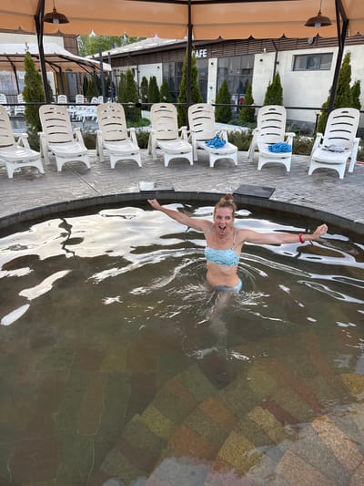 A woman in a light blue bikini is in a hot spring surrounded by a stone border and filled with water. She has her arms raised in a joyful manner, and is wearing a red bracelet on her left wrist. The hot spring is located outdoors. To the left of the pool, there is a patio area with several white plastic chairs arranged in rows under a covered awning, indicating a cafe or resort setting. The patio is lined with trees. In the background, there is a light-colored two-story building with windows and some greenery. The image was taken in Kara-Oy, Kyrgyzstan.