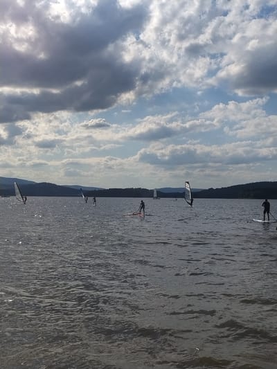 The image depicts a wide, calm lake with a hilly, tree-covered shoreline in the distance. The sky is filled with a mixture of dark, dramatic clouds and bright, lighter ones, suggesting dynamic weather conditions. Several people are engaged in water sports on the lake. On the left side, a person is windsurfing with a white sail. Further into the lake, two other individuals are visible on what appear to be stand-up paddleboards. Towards the right side of the frame, another person is stand-up paddleboarding, and another is windsurfing. The water is choppy with ripples, and sunlight is glinting off the surface in several areas. The scene is likely set at the Radslav Reservoir, a popular recreational area near Radslav in the Czech Republic, given the landscape and activities.