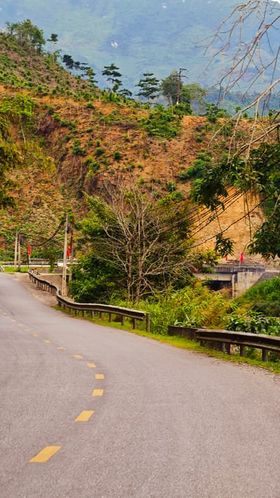 The scene is a road winding through a mountainous area in Minh Tân Commune, Vietnam. The road, marked with dashed yellow lines, is lined with a metal guardrail on the right side. A bridge spans a small valley, with red flags visible on either side. The landscape is characterized by steep, eroded hillsides covered in sparse vegetation and some trees. Electrical poles and wires run alongside the road, adding to the infrastructure. The backdrop features distant mountains shrouded in mist, contributing to the natural setting of the photograph.
