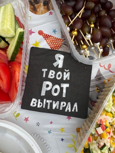 The image shows a close-up, overhead view of a table spread, likely for a casual gathering or party. There are several plastic containers holding food items. On the left, a container holds sliced cucumbers and tomatoes. In the center, a black, textured napkin or placemat displays white text in Russian, which reads "Я твой РОТ ВЫТИРАЛ" (meaning "I wiped your MOUTH"). To the right of the napkin, a container holds grapes on skewers, some decorated with gold and pearl-like beads. Further to the right, another container holds a salad composed of corn, chopped imitation crab meat, and what appears to be cucumber and other ingredients. The table is covered with a white disposable tablecloth featuring a festive pattern of stars, party hats, and cartoon cat illustrations. The overall scene is brightly lit, suggesting an indoor or shaded outdoor setting. No specific location in Valok, Russia can be determined from the image alone.