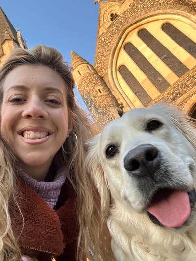 An outdoor close-up image features one adult female and one light-colored Golden Retriever dog positioned in front of St. Thomas of Canterbury Church in Brentwood, United Kingdom. The female, with blonde hair, smiles directly at the camera, displaying teeth, and wears a rust-colored faux-shearling jacket over a light purple ribbed turtleneck, with silver hoop earrings visible. The dog, situated to her right, also looks towards the camera, with its mouth slightly open and tongue partially extended. The background consists of the stone facade of St. Thomas of Canterbury Church, showcasing a large arched window with dark panes, intricate stone carvings, and two stone spires or turrets, set against a clear blue sky. The scene is brightly lit by natural sunlight, highlighting the warm tones of the church stone.