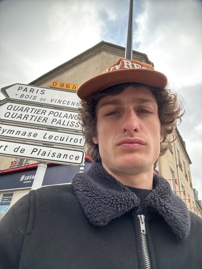 The image depicts a male individual in a close-up, chest-up outdoor portrait, set against an urban background in Joinville-le-Pont, France.
The subject is a young adult male with medium-length, curly brown hair. He wears an orange-brown baseball cap with white text, partially visible as "Ulv Res," worn forward. His facial features include a straight nose with a small dark spot on the right side, and full lips, with a neutral expression as he looks directly into the camera. He is dressed in a black jacket or coat featuring a dark grey, textured, sherpa-style collar and a visible silver zipper running down the front. A dark, possibly black, crew-neck garment is visible underneath the jacket.
Behind the individual, a white street signpost points to the left, displaying multiple destinations: "D 86 b" (a road designation), "PARIS - BOIS DE VINCENNES," "QUARTIER POLANGIS," "QUARTIER PALISSY," "Gymnase Lecuirot," and "Port de Plaisance." These signs identify the location as Joinville-le-Pont, a commune in the southeastern suburbs of Paris, France. Below the signs, a blue and white storefront sign for "EIFFAGE IMMOBILIER" is partially visible.
In the background, to the left, a light-colored building with architectural details like a cornice is visible. To the right of the subject, another multi-story, light-colored building with numerous windows and some shutters is present. A tall, slender, dark-colored pole extends vertically from behind the subject's head towards the top of the frame. The sky is overcast with light grey clouds.