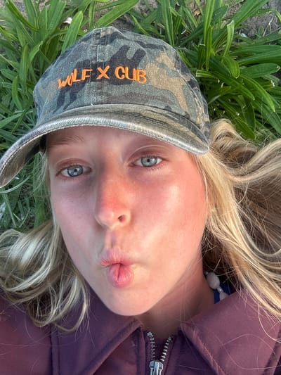 A young Caucasian woman is lying on her back in what appears to be a grassy area. She is wearing a camouflage cap with "WOLFX CLUB" written in orange on the side, and a purple jacket. She has blonde hair and light-colored eyes. Her lips are pursed in a kissing gesture. A necklace with white beads can be seen. The background appears to be green grass or other vegetation. It's impossible to determine the exact location within the Wollongong City Council area without more context or visual cues.
