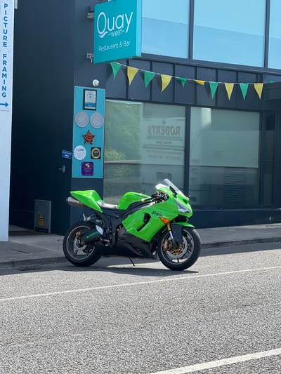 A bright green Kawasaki Ninja motorcycle is parked on a street in front of a building. The building has a dark blue facade and a sign that reads "Quay -west- Restaurant & Bar". There are colorful triangular flags strung across the front of the building. On the wall of the building are several awards and certifications. To the left of the motorcycle, a white sign with blue text reads "PICTURE FRAMING". The motorcycle is positioned on the street, with its front wheel slightly turned. The street is paved with asphalt and has a white line painted on it. The background consists of the building and its windows, reflecting the sky.