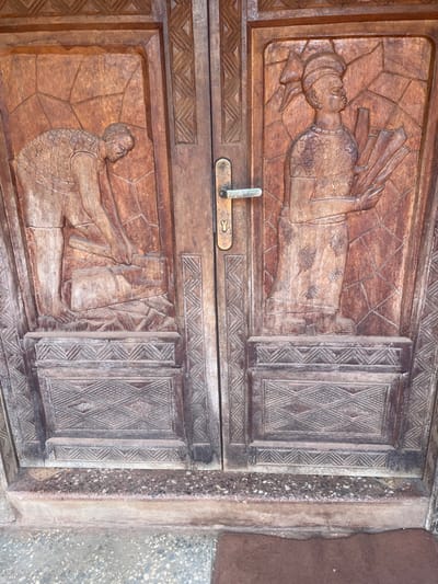 A wooden door displays intricate carvings of human figures. On the upper panel, a person lies horizontally with outstretched arms holding a fan-like object. The lower panel depicts another figure reaching for a vase. Each panel has adjacent sections with geometric diamond patterns. A simple metal handle and lock are centered between the two carved scenes. The door is weathered, and its surface shows signs of age and wear.