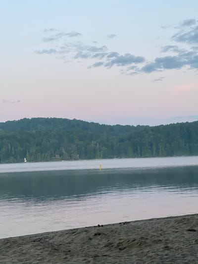 This is a photograph of a serene lake scene, likely at dawn or dusk, with a sandy beach in the foreground. The water is calm, with a gentle mist rising from its surface. Beyond the lake, a dense forest covers a rolling hillside. The sky is a soft gradient of pale blue and pink, with a few wispy clouds. A small yellow buoy is visible in the water, and a sailboat with a white sail can be seen near the shore on the left side of the image. The beach is covered in sand, with some scattered debris. The overall atmosphere is peaceful and natural. This location is likely in Duhamel, Canada, given the natural landscape and typical weather conditions for the region.
