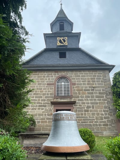The image depicts the exterior of a stone church in Asterode, Germany. The church features a dark gray slate roof, a prominent clock tower with a golden clock face, and an arched window above the entrance. In front of the church is a large, silver bell with an inscription. The foreground is filled with green bushes and grass, and to the left, there are trees. A stone bench is visible behind the bell. The sky is overcast.