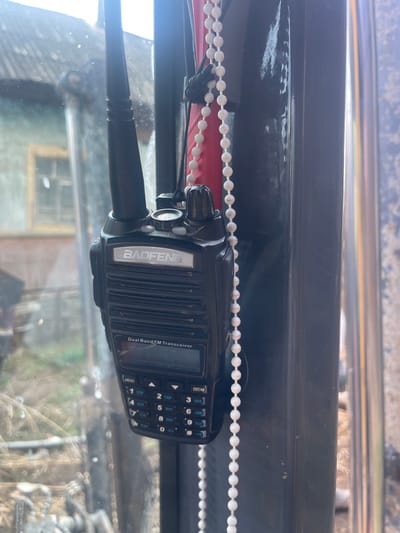 A Baofeng Dual Band FM Transceiver is mounted inside a vehicle, likely an excavator or heavy machinery due to the rugged environment visible through the windshield. The radio is positioned near a white beaded chain. Outside, the environment suggests a rural or industrial location in the city of Поедуги, Russia. Visible through the glass is an indistinct building, and the surroundings appear to be slightly unkempt with what seems to be foliage or brush.