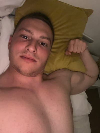 A young man is depicted lying down, shirtless, in what appears to be a bed. He is positioned on his back, looking directly towards the camera with a neutral and calm expression. His head rests on a white pillow, with a mustard-yellow pillow placed next to it. His right arm is bent at the elbow, with his fist lightly clenched, resting on the yellow pillow. The man has short, dark hair and visible stubble on his chin and cheeks. The lighting in the image is dim, suggesting an indoor setting like a bedroom, possibly during the evening or night. White bedding and additional pillows are visible around him. His bare torso and shoulders are visible. No specific location cues are present within the image itself, but the provided context is Vitry-sur-Seine, France. There is no visible text in the media file.