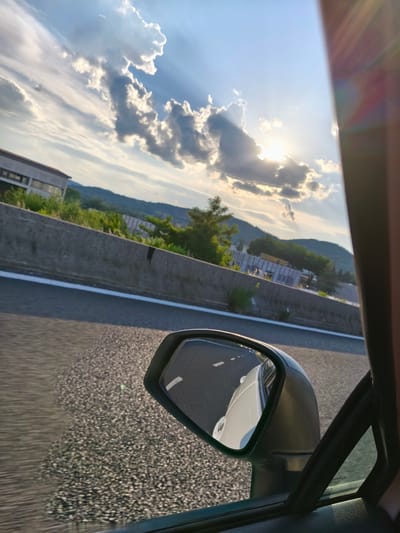 This media file captures a view from a car traveling on a highway, likely in San Pietro, Italy. The time of day is late afternoon or early evening, as indicated by the low angle of the sun and the dramatic crepuscular rays piercing through the clouds. The weather appears clear with scattered cumulus clouds, creating a picturesque sky.

The primary subjects include the road, a concrete barrier, distant hills covered in greenery, and a building with windows. The foreground is dominated by the car's side mirror, reflecting the road and the car's white exterior. The side of the car's interior, including a window and the frame of the door, are also visible.

The activity is clearly a journey by car. The motion blur on the road surface suggests the vehicle is in motion. The reflection in the side mirror, showing the lane markings and the white car, further reinforces the context of travel.

Notable details include the texture of the asphalt road, the rough surface of the concrete barrier, and the lush vegetation on the hills. The sun's rays create a lens flare effect on the right side of the frame, adding to the dramatic lighting. The scene evokes a sense of travel and perhaps peaceful contemplation of the scenery passing by.

There is some faint text visible on the building in the distance, but it is too blurry to be deciphered. The overall mood is serene and visually striking due to the interplay of light and shadow in the sky and on the landscape.