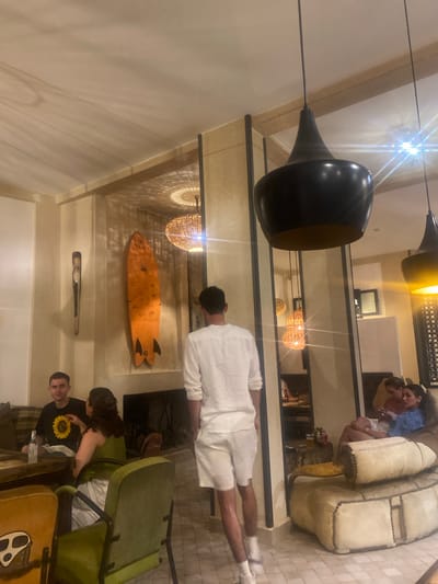In the city of Taghazout, Morocco, an indoor scene unfolds with a group of people gathered in a dimly lit, tastefully decorated establishment. The ambiance suggests a relaxed social gathering, possibly a café or lounge, characterized by warm lighting and contemporary decor.
A prominent feature of the space is the wall adorned with a vibrant orange surfboard, a nod to Taghazout's surfing culture. Adjacent to it hangs an intriguing wooden artifact, adding an artistic touch to the environment. Overhead, a large, modern black pendant light fixture dominates the upper portion of the frame, its sleek design contrasting with the woven texture of smaller pendant lights visible in the background. These smaller lights cast intricate patterns on the ceiling, enhancing the warm and inviting atmosphere.
Several individuals are present, engaged in various activities. In the foreground, a young man in a white shirt and shorts walks away from the camera, his posture suggesting movement through the space. To the left, two individuals are seated at a table. One man, wearing a black t-shirt with a sunflower print, appears to be conversing, while a woman beside him, dressed in a green top, actively engages in conversation, her hand gesturing. Further to the right, a couple is seated on a plush, cream-colored couch. They appear relaxed, with the woman in a blue top looking towards the camera, and the man beside her looking down.
The seating arrangements include a mix of chairs and sofas, with a striking green armchair positioned prominently in the foreground. The flooring is a light-colored tile, contributing to the overall bright yet cozy feel of the establishment. The background reveals more seating areas and decorative elements, including what appears to be a fireplace, further immersing the viewer in the tranquil, social environment of Taghazout.