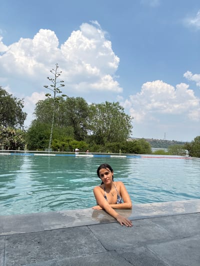 A woman with dark hair and wearing a patterned bikini is in an outdoor pool. She is leaning against the grey stone edge of the pool. The pool is lined with blue and white lane markers. Behind the pool is a green landscape with trees and a tall flowering plant, set against a blue sky with large white clouds. In the distance, a hill with a transmission tower on top is visible. This photograph was taken at Pueblo Nuevo de Jasso, Mexico.
