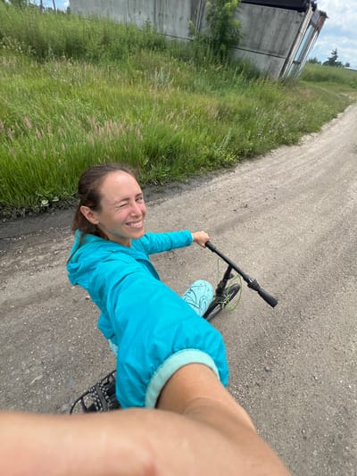 A person is riding a bicycle on a dirt road in Pivkino, Russia. The person is wearing a bright blue hooded jacket and light blue patterned leggings. They are smiling and appear to be enjoying themselves. The road is surrounded by tall green grass and some small wildflowers. To the left of the road, there is a building with a concrete wall and some windows. The sky is cloudy, suggesting it might be an overcast day. The image is taken from a first-person perspective, as if the viewer is the one riding the bike. There is no visible text in the image.