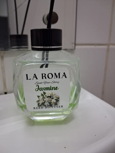 This image shows a clear glass reed diffuser with a light green liquid inside. The diffuser has a black cap and is placed on a white surface, possibly a sink or countertop, in what appears to be a bathroom. The background consists of tiled walls. A reflection of the diffuser and surrounding environment is visible on the white surface.
The label on the diffuser features text and an illustration. The text includes "LA ROMA," "Scent Your Story," "Jasmine," and "REED DIFFUSER." The illustration depicts a cluster of white jasmine flowers with green leaves.
The overall impression is one of a domestic setting, likely a bathroom in Constanța, Romania, given the location context. No people are visible, and the focus is on the product. The lighting suggests an indoor environment, possibly daytime, though no specific time or weather cues are apparent. The image is well-lit, highlighting the details of the diffuser and its label.