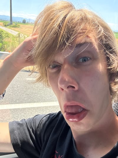 This media file is a close-up, selfie-style portrait of a young person with blonde hair, taken outdoors. The individual's face is the primary subject, tilted towards the camera. They have blue eyes and are sticking their tongue out slightly, with their mouth open. Their blonde hair is tousled and falls across their forehead, obscuring part of their face.

The background suggests a rural or semi-rural setting. On the left, a field with yellowing crops and green foliage is visible under a bright blue sky, indicating it is daytime and likely fair weather. A road or path runs through the landscape, and some distant mountains can be seen. On the right, a textured, light-colored surface, possibly a wall or railing, is present. The overall lighting suggests it is a sunny day.

The individual is wearing a dark t-shirt. On their left wrist, a bracelet with silver charms is visible. The composition is casual and playful, with the subject making a facial expression. The orientation of the image is rotated, as if taken by someone holding the camera while looking down.

The provided location context is Cadeo, Italy. There is no visible text in the image.